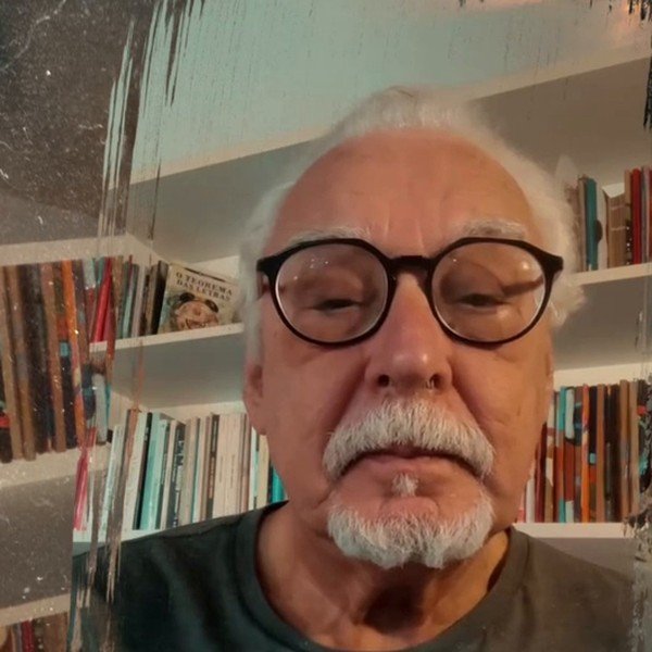 Carlos Pessoa Rosa, de cabelos e barba brancos e óculos de armação escura, lendo o poema Chuva diante de uma estante de livros