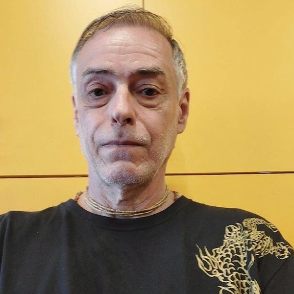 Cláudio Daniel, de cabelos grisalhos e camiseta preta com um dragão dourado, diante de uma parede amarela