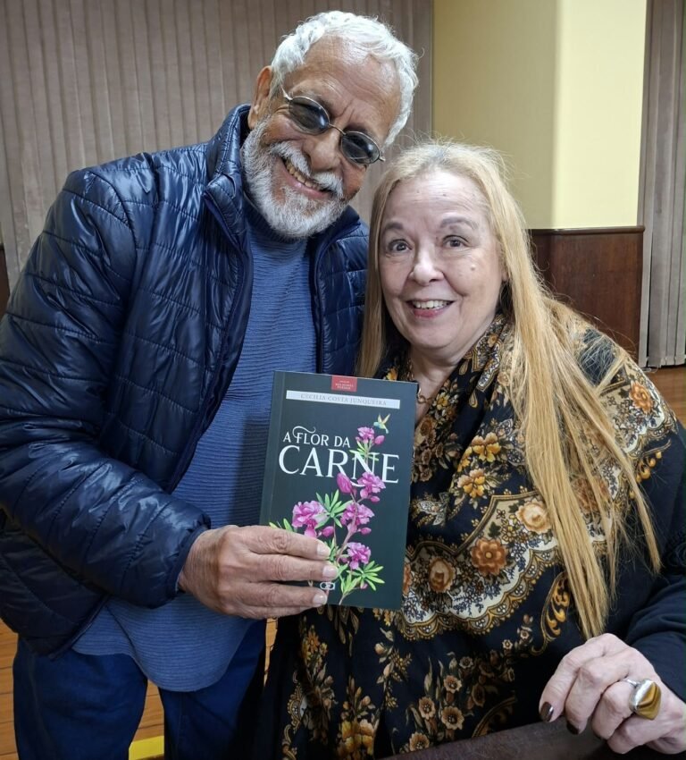 Tanussi Cardoso, de barba branca e óculos, ao lado de Cecilia Costa Junqueira, os dois sorrindo e segurando o livro A flor da carne